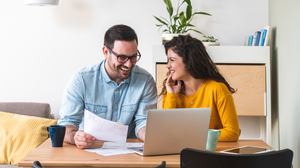 A man and a women with a laptop computer. The lady is looking at the man and the man is smiling and holding a sheet of paper