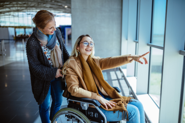 Person in wheelchair pointing out of window. Carer holding chair
