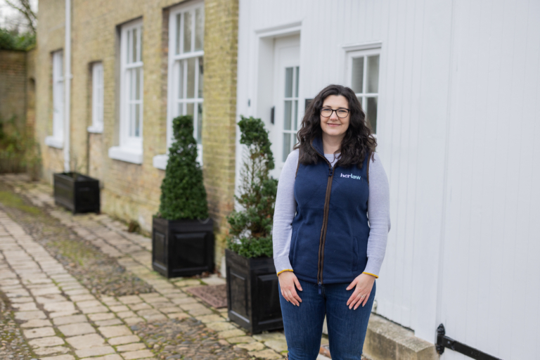 lady with long dark brown hair and glasses standing in front of a house with jeans and a blue gilet on.