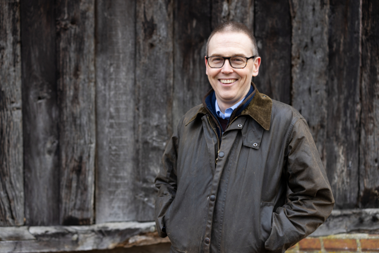 Man smiling wearing a green jacket standing in front of a brown wooden building. His hand are in his coat pockets and he is wearing glasses