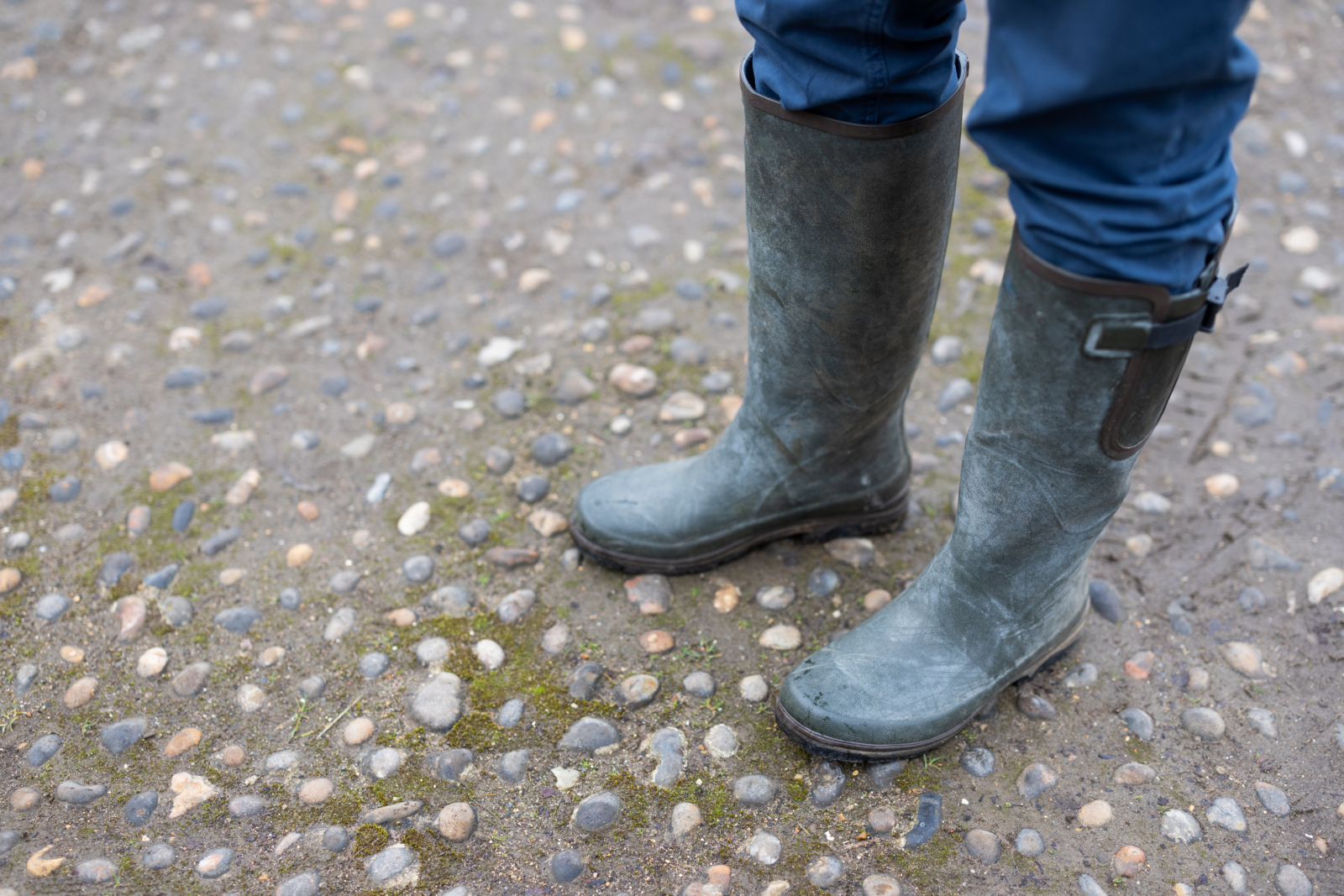 image shows a person standing with green wellingtons, you cannot see their head, the photo is of their feet only. The person is standing on grey and white cobble stones. Some are covered in green moss
