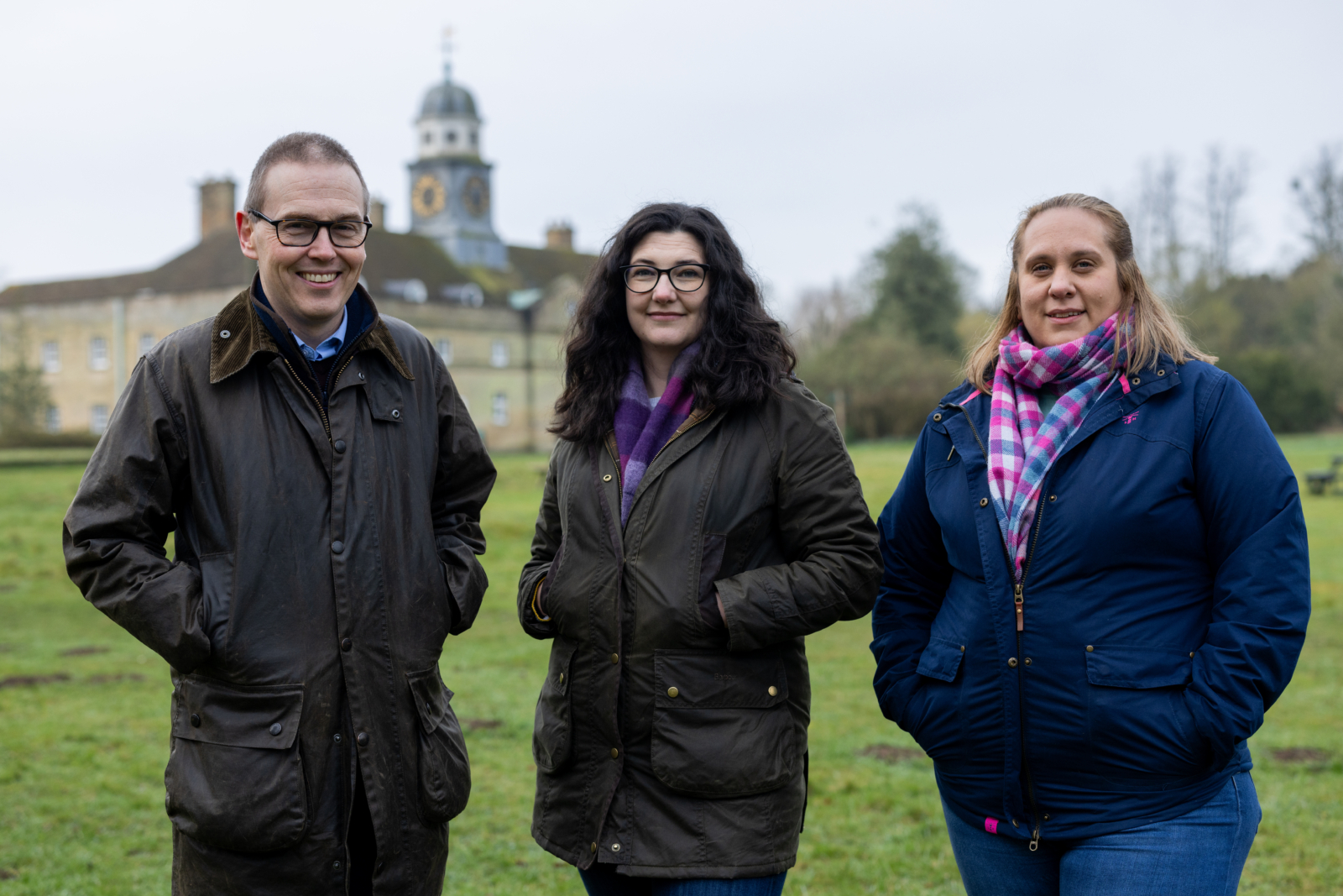 two women and one man standing in a field with a stately home in the background