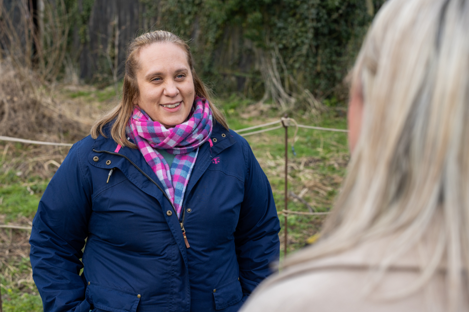 A lady with a blue coat and pink and purple scarf smiling and facing a lady with blonde hair. You can only see the back of the head of the lady with blonde hair. They are having a conversation and smiling. Behind the lady with the blue jacket is a rope fence and some tree with leaves on the ground