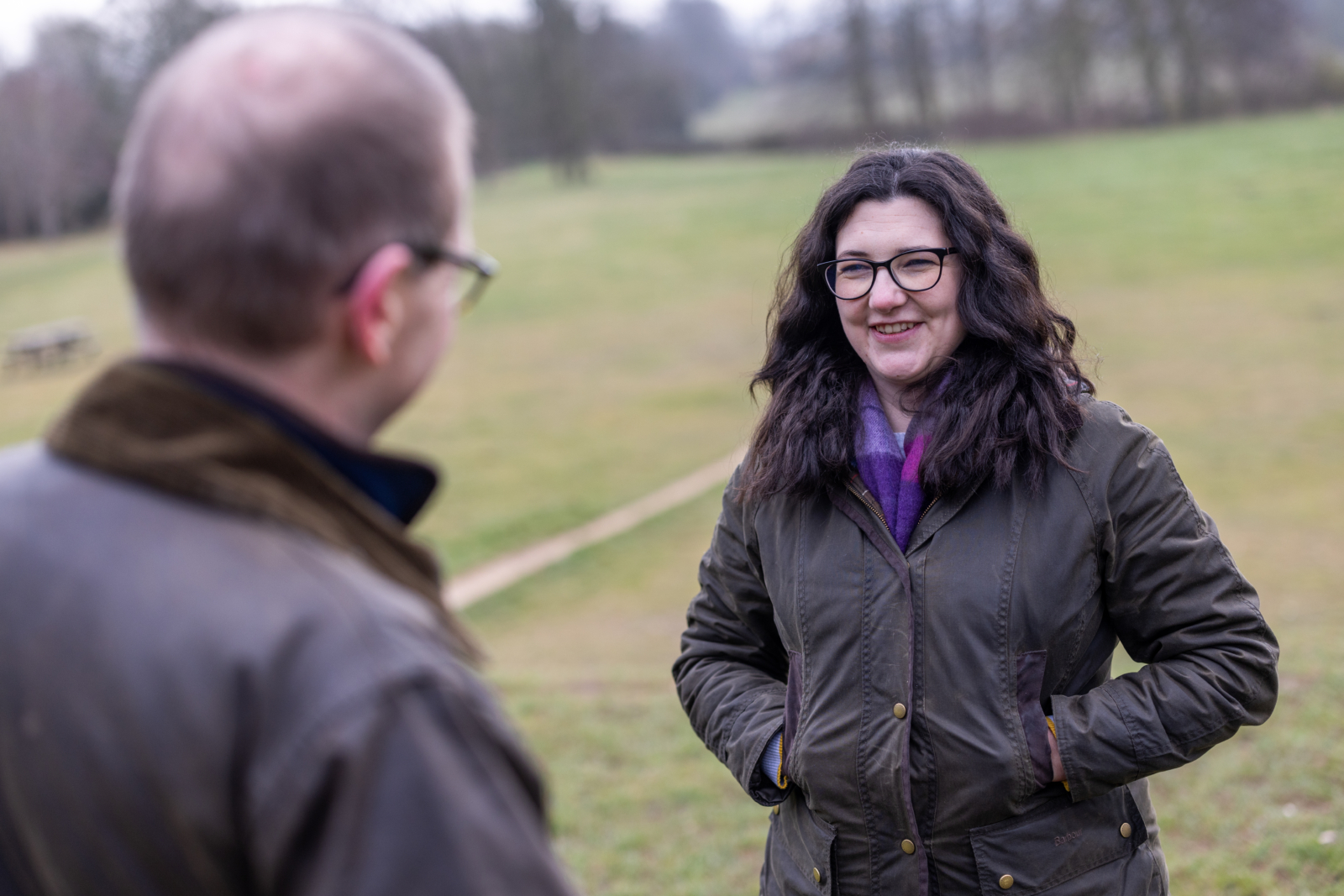 two people standing in a field, one man and one woman. Both wearing green coats. The lady has her hands in her pockets and is smiling at the man. They are having a conversation