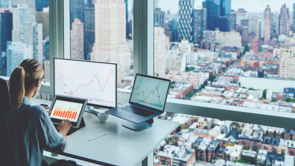 Person at a desk with laptop and screens in front of large window with city view