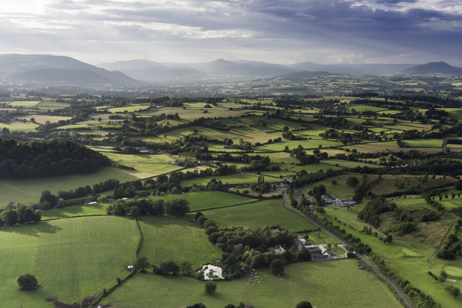 Rays of sunlight illuminating the picturesque patchwork quilt landscape of green fields, farms and misty mountains under panoramic skies from high in a hot air balloon. ProPhoto RGB profile for maximum color fidelity and gamut.
