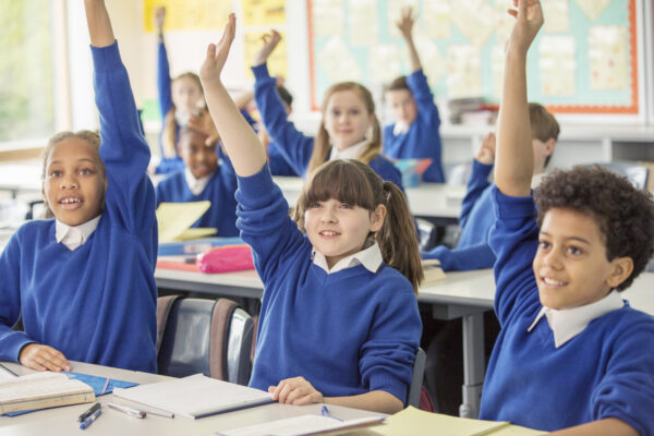 Primary school children wearing blue school uniforms raising hands in classroom