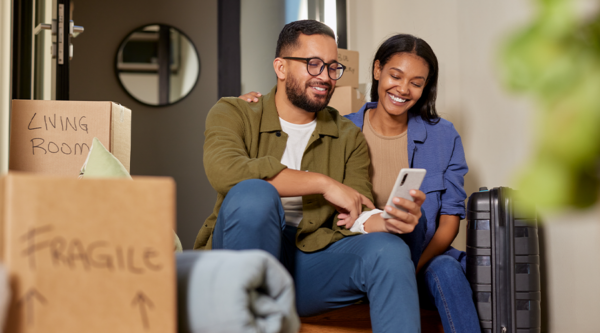 A smiling couple sits on stairs surrounded by moving boxes, looking at a smartphone.