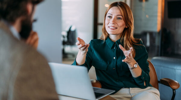 Person seated in an office meeting room gesturing while speaking across a table with an open laptop, engaged in a professional discussion with another partially visible individual.