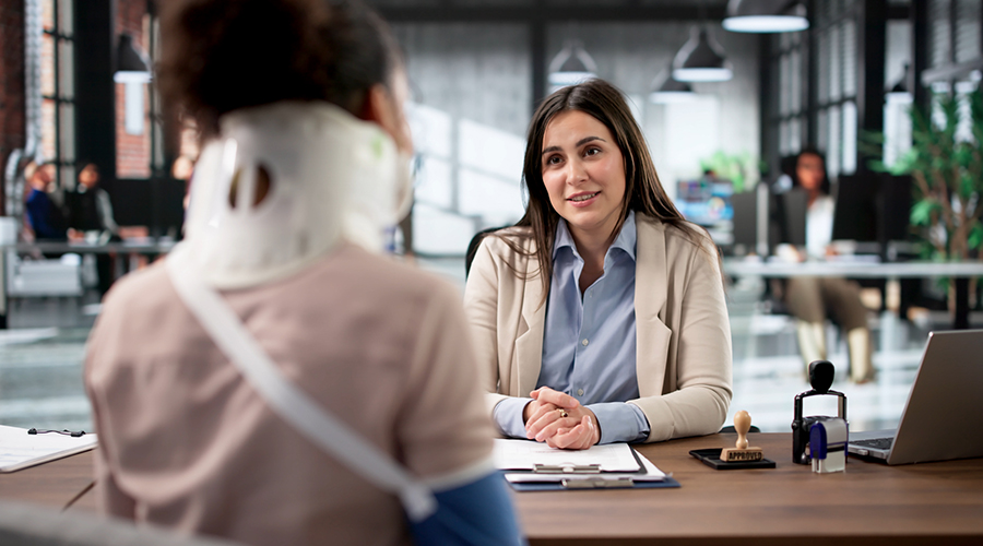 A consultation in a modern office where an individual wearing a neck brace sits across a desk from another person, with documents and a laptop visible, suggesting a discussion about a potential injury claim.