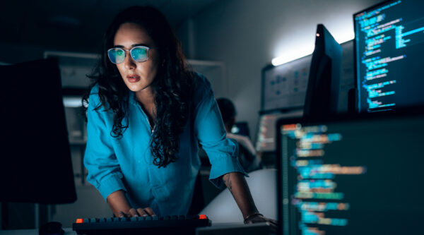 An individual working at a computer in a low‑lit office, with multiple monitors displaying lines of code, suggesting activity related to data processing or information security.