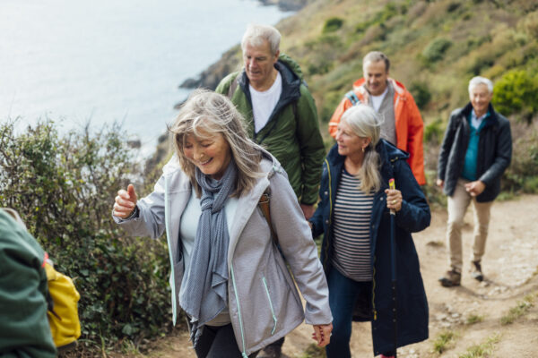 A group of five adults walking along a coastal path, wearing outdoor jackets and carrying backpacks. One person in the foreground leads the group, while others follow in a line behind. The path runs alongside a hillside with greenery, with the sea visible below on the left.