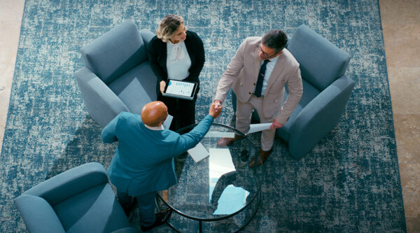 Overhead view of three professionals seated around a round glass table in a meeting area, with two individuals shaking hands while another holds a tablet displaying data, suggesting a business agreement or corporate negotiation.
