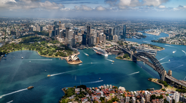 Aerial view of Sydney Harbour featuring the Sydney Opera House, Harbour Bridge, waterfront skyline and boats on blue water under a partly cloudy sky.