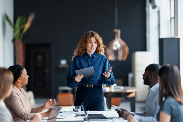 A woman holding a tablet leads a workplace discussion with colleagues seated around a meeting table.