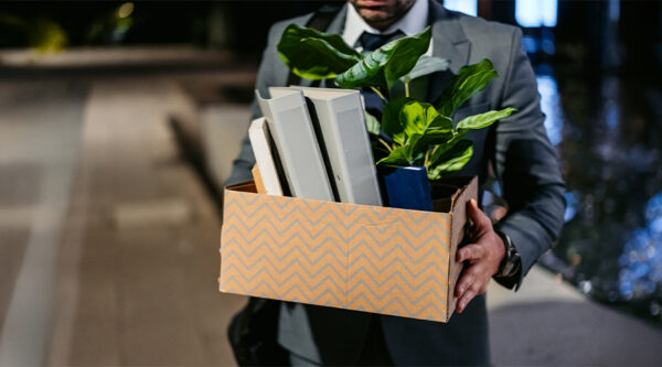 Person in business attire carrying a box of office belongings, including folders and a potted plant.