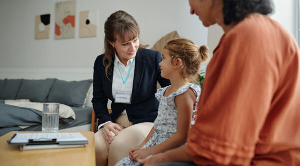 Social worker speaking with a child during a home visit while another adult sits nearby.