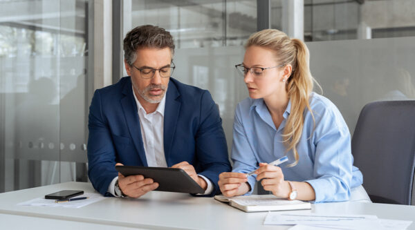 Two professionals seated at a desk in a modern office, reviewing information on a tablet together.