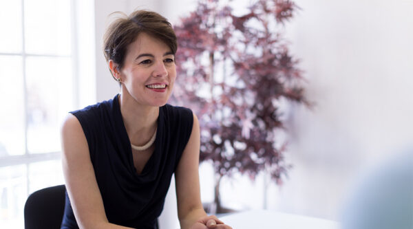 Professional seated at a table in a bright office, with a blurred potted tree in the background.