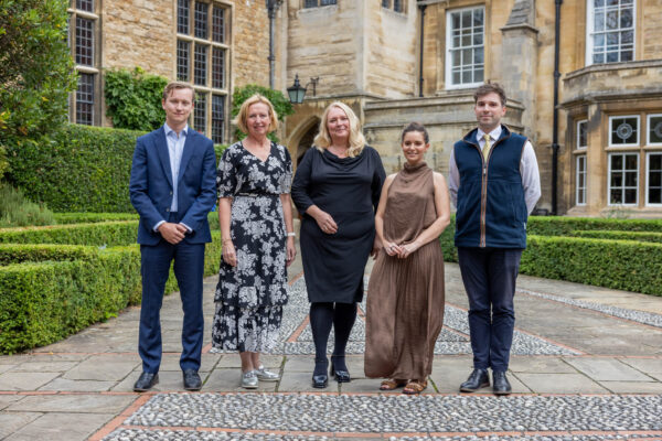 Five people stand in a formal landscaped courtyard in front of a historic stone building with large windows, trimmed hedges and decorative paving.