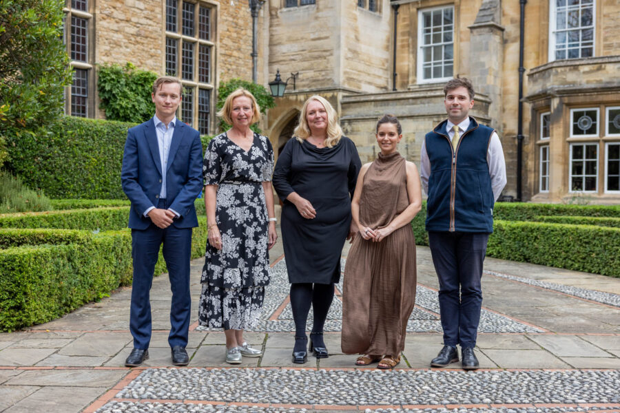 Five people stand in a formal landscaped courtyard in front of a historic stone building with large windows, trimmed hedges and decorative paving.