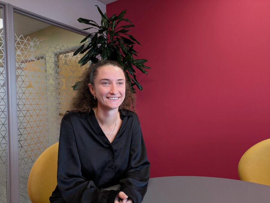 Person wearing a black blouse seated at a round table in a modern office meeting area, with a red feature wall, frosted glass partition, yellow chairs, and a large green plant in the background.