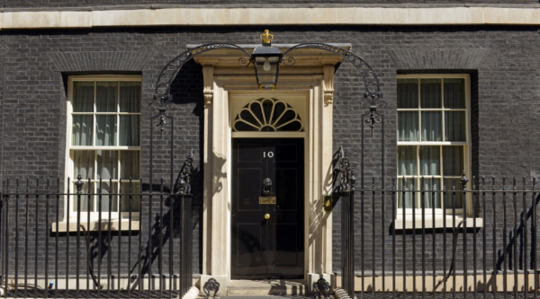 The black front door of 10 Downing Street, framed by cream stonework, with an overhead lantern and black iron railings.