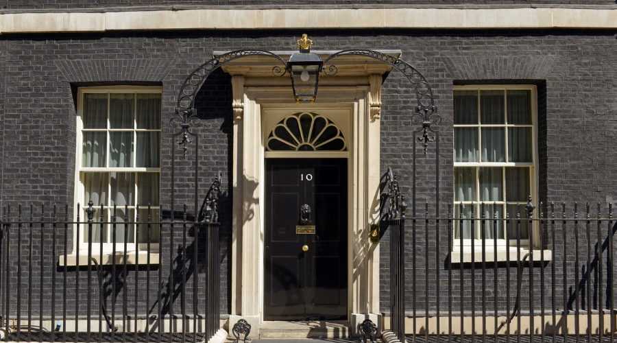 The black front door of 10 Downing Street, framed by cream stonework, with an overhead lantern and black iron railings.
