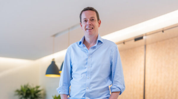 Person standing indoors in a modern office environment, wearing a light blue button-down shirt.