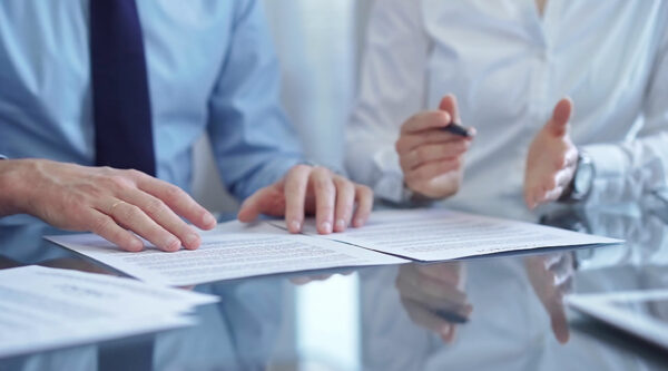 Two professionals reviewing contract documents at a desk, with one pointing to the text and the other gesturing with a pen.