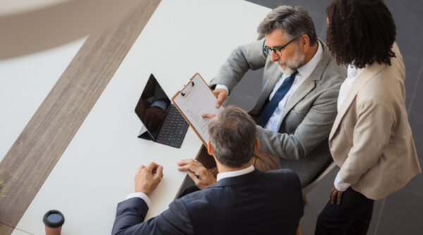 Three professionals review a contract on a clipboard during a meeting at a desk with a laptop.