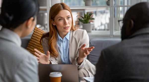 Three people sit around a laptop in a modern office. The person facing the camera gestures with both hands during a discussion, with a takeaway coffee cup and papers on the table.