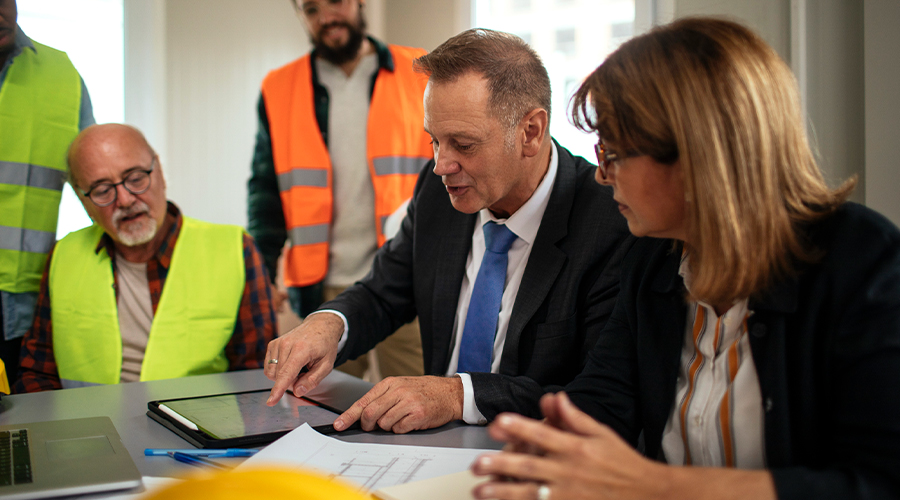 Construction professionals review project plans and payment details on a tablet during a meeting.