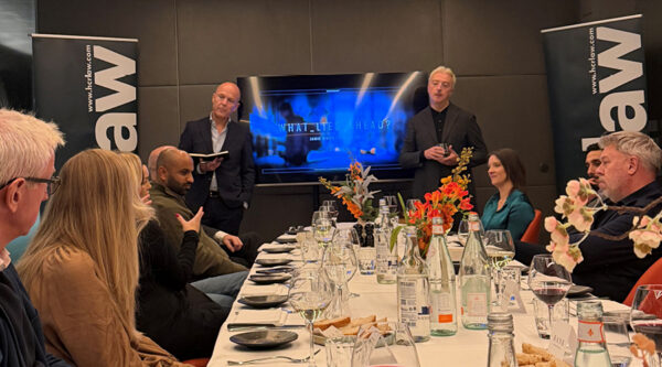 Attendees seated around a long, formally set dining table during a business event. Two speakers stand at the front of the room beside a large screen displaying the title “What Lies Ahead?” and branded banners.