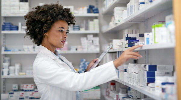 Person in a white coat using a tablet while checking medicine stock on pharmacy shelves.