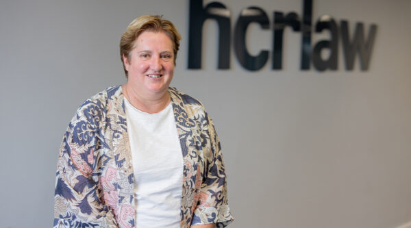 Person in a patterned jacket standing in front of the HCR Law logo.