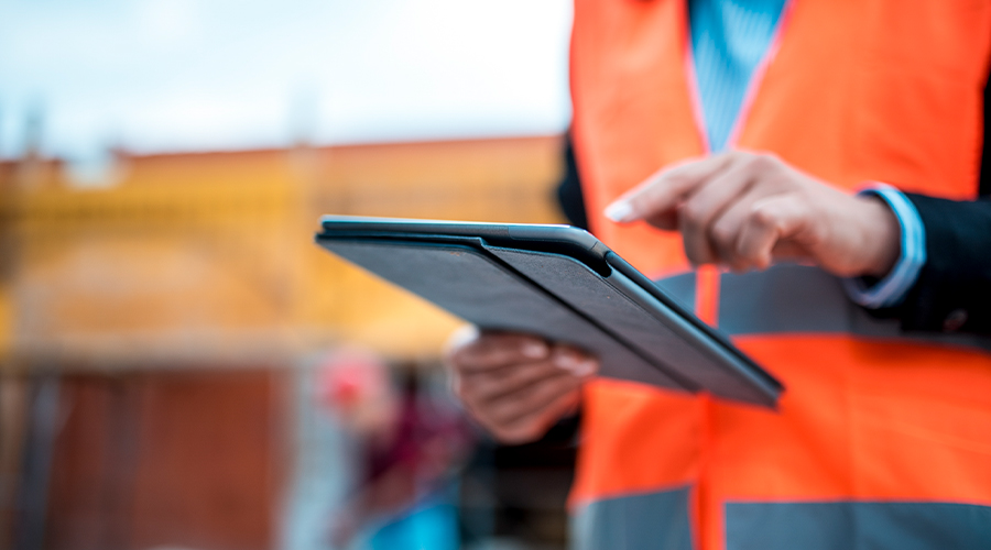 Close-up of a person wearing a high-visibility orange safety vest using a tablet on a construction site, with a blurred building project visible in the background.