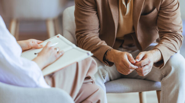 A person takes notes while speaking with another person seated opposite in a consultation.