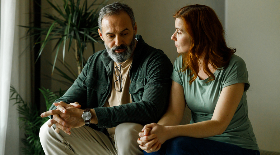 Two adults sit closely together indoors; one looks down with hands clasped while the other turns towards them, with houseplants in the background.