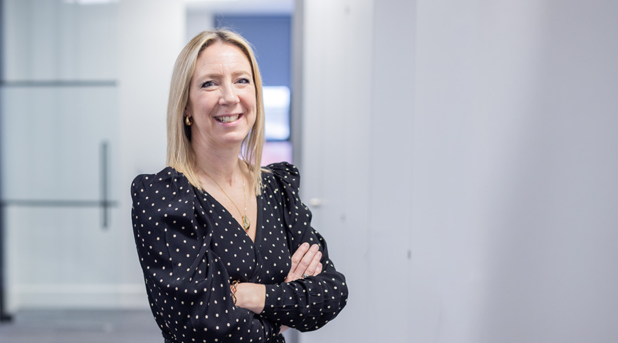 Professional portrait of a person in a black polka-dot blouse, standing with arms crossed in a modern office.