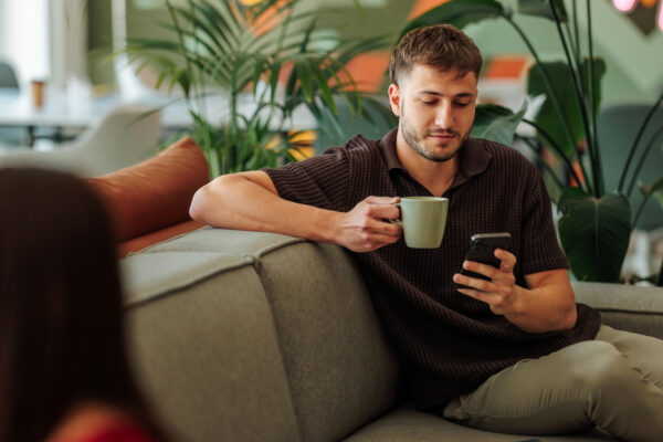Young adult employee enjoying a coffee break, using his smart phone on a comfortable sofa in a modern office environment
