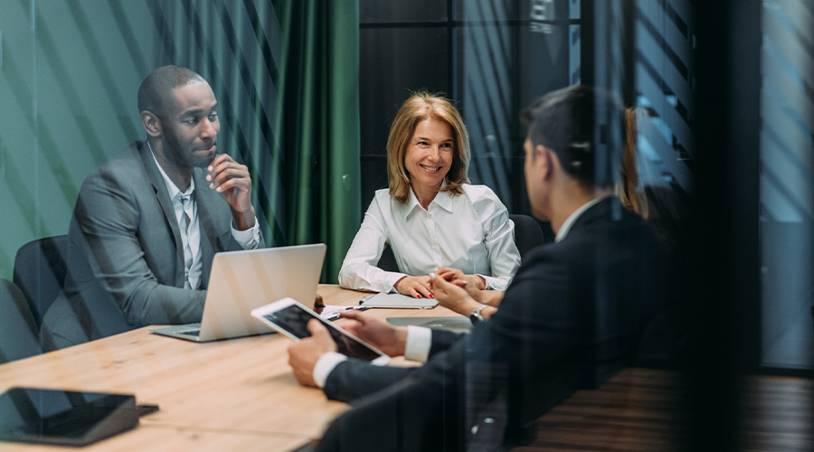 Three professionals discussing work around a meeting table with a laptop and tablet in a modern office.