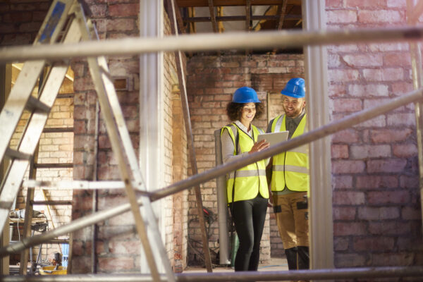 Two construction professionals in hard hats and high-visibility vests reviewing a tablet at a building renovation site.