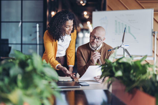 Two colleagues review documents beside a model wind turbine and sustainability charts in a plant-filled office.