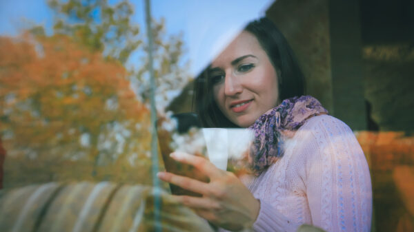 Person using a tablet while holding a mug, seen through a window reflecting autumn trees.