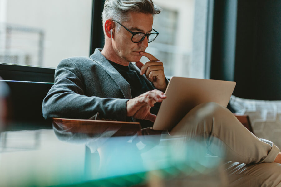 Person wearing glasses and an earbud, seated by a window and using a laptop.