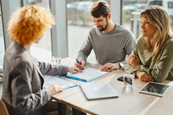 Person signing documents during a meeting with two other people seated across a table in a modern office.