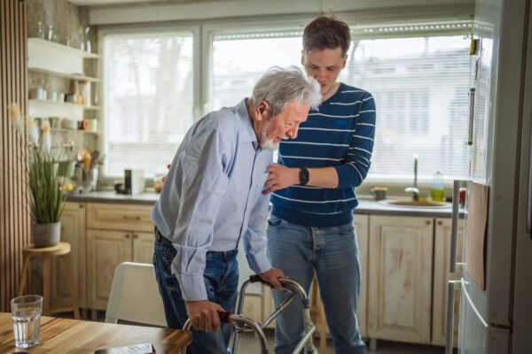 Two adults in a bright kitchen, one using a walking frame while the other stands close behind with a hand positioned to provide physical support.