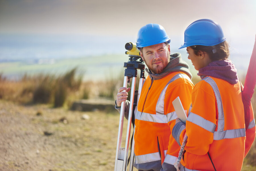 Two construction professionals wearing high-visibility orange jackets and blue safety helmets stand outdoors beside surveying equipment on a tripod, discussing site measurements in a rural landscape.